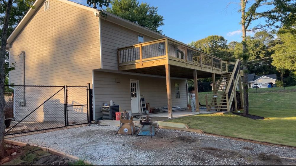 Two-story house with a wooden deck and stairs. Gravel driveway and a grassy yard.