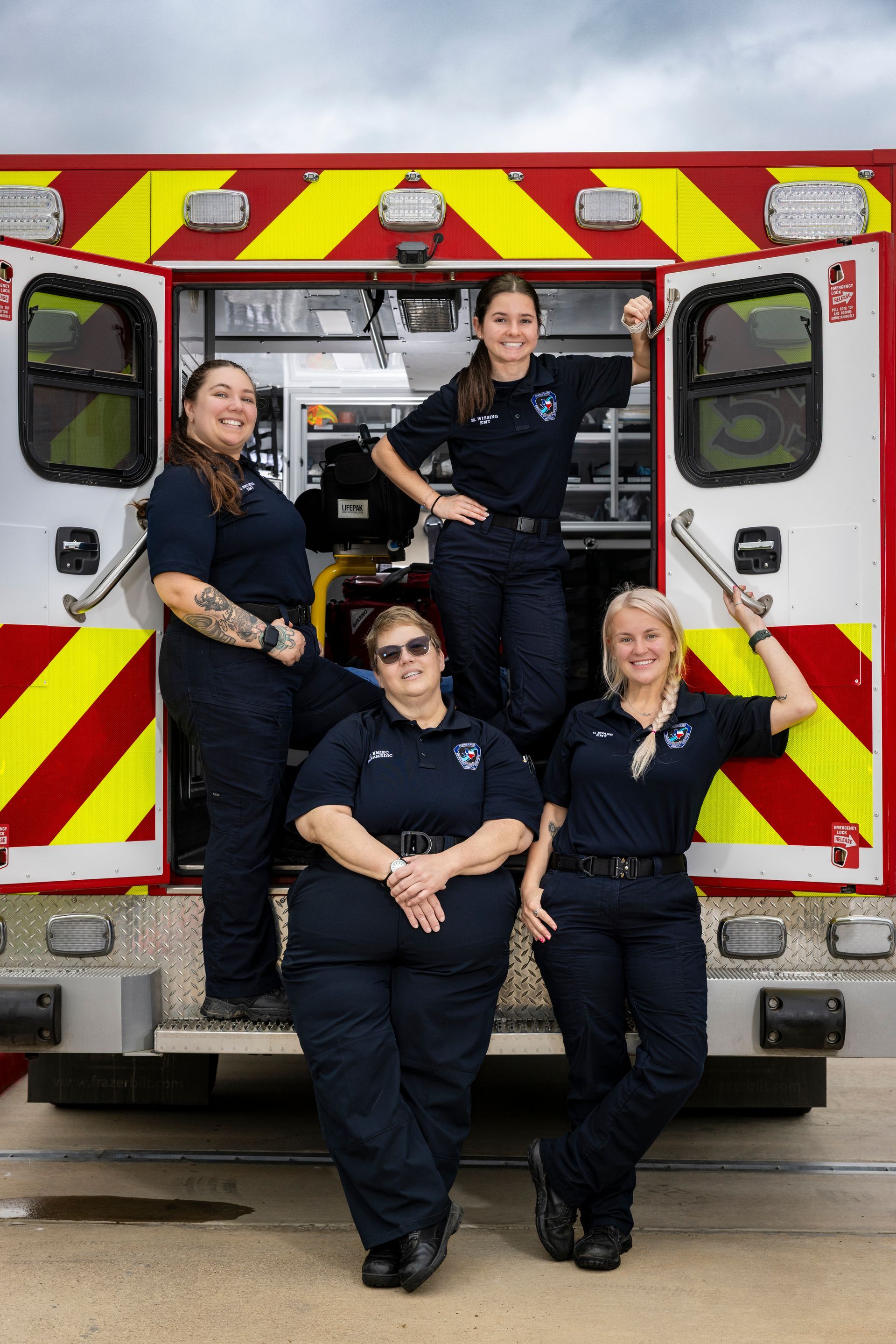 Four firefighters posing at the open back of a red-and-yellow ambulance truck.