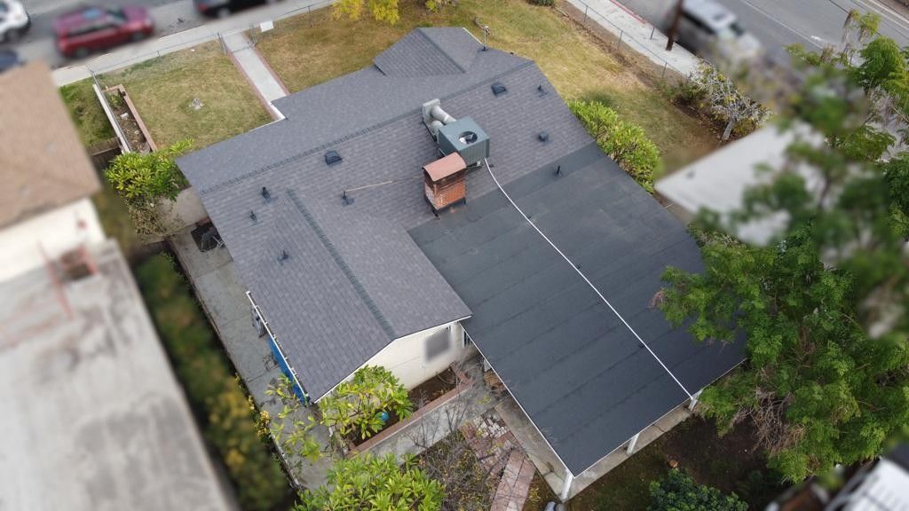 Overhead view of a house with a dark gray roof, an AC unit, and nearby trees and a street.