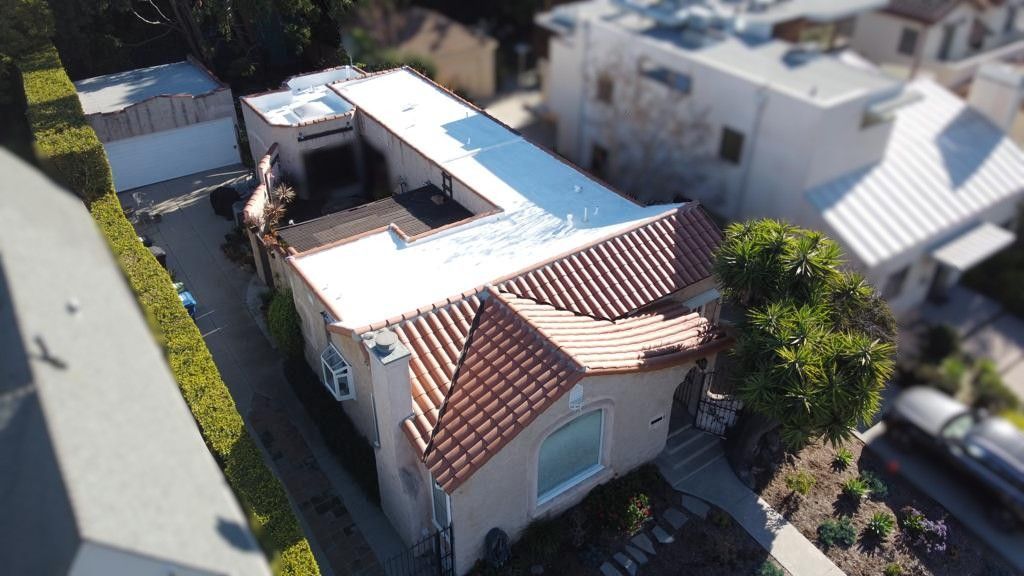 Aerial view of a light-colored stucco house with red-tiled roof, surrounded by greenery and other houses.