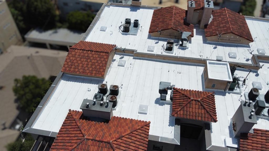 Overhead view of a flat roof with red tile roofs, vents, and air conditioning units. White roof sections.