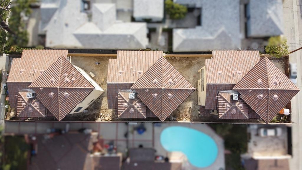 Overhead view of three houses with brown tile roofs, a swimming pool below, and surrounding houses.