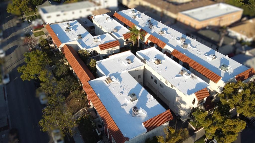 Aerial view of a cream-colored apartment complex with red tile roofs and mature trees.