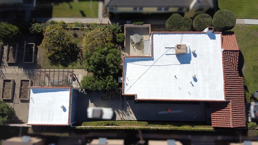 Overhead view of a house with a white roof, garden beds, and surrounding trees.