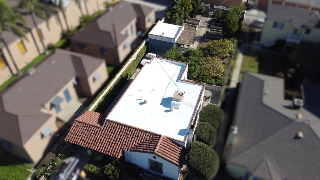 Aerial view of homes in a neighborhood; houses with brown roofs, green trees, and white or light-colored walls.