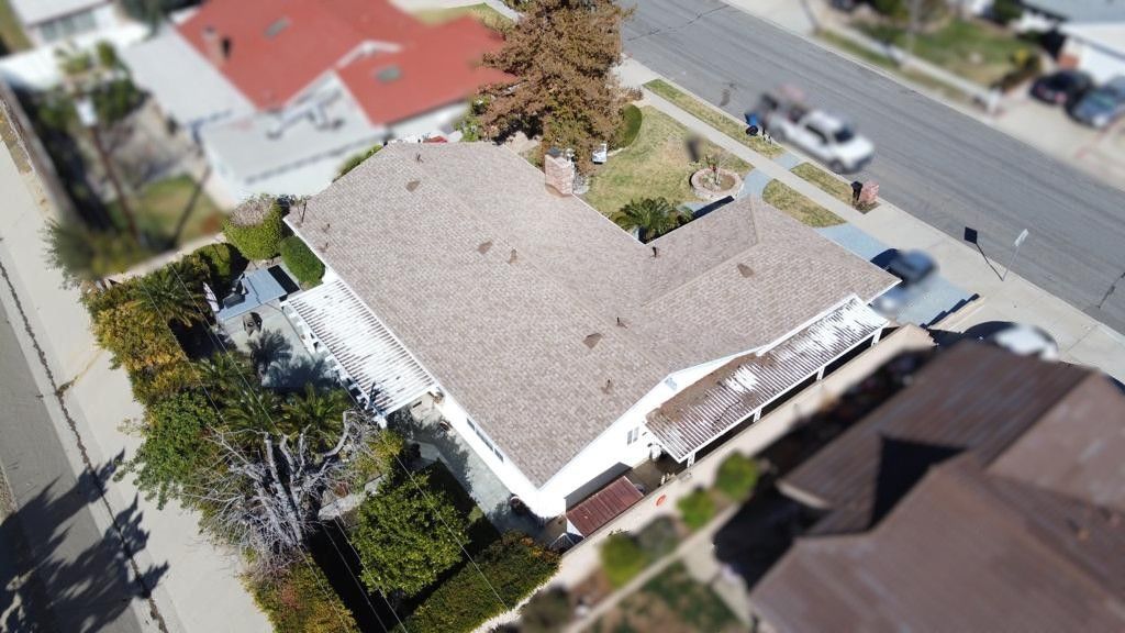 Overhead view of a white house with a brown roof, surrounded by trees and bushes, street in background.