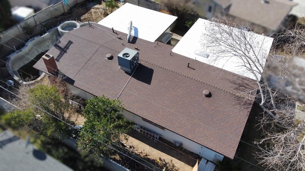 Overhead view of a brown-roofed house with a white flat roof, surrounded by trees and a fence.