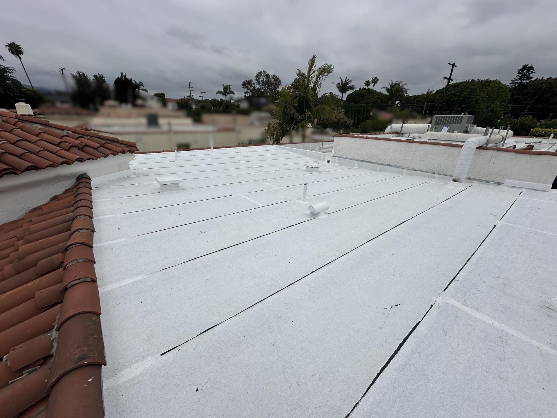 Flat white roof with visible seams, vents, and a section of red tile roof. Overcast sky.