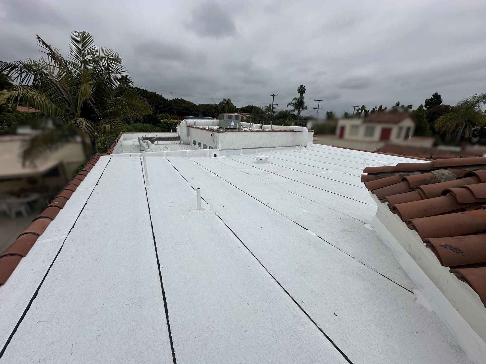 A white flat roof with black seams, surrounded by red tile roof and a cloudy sky.