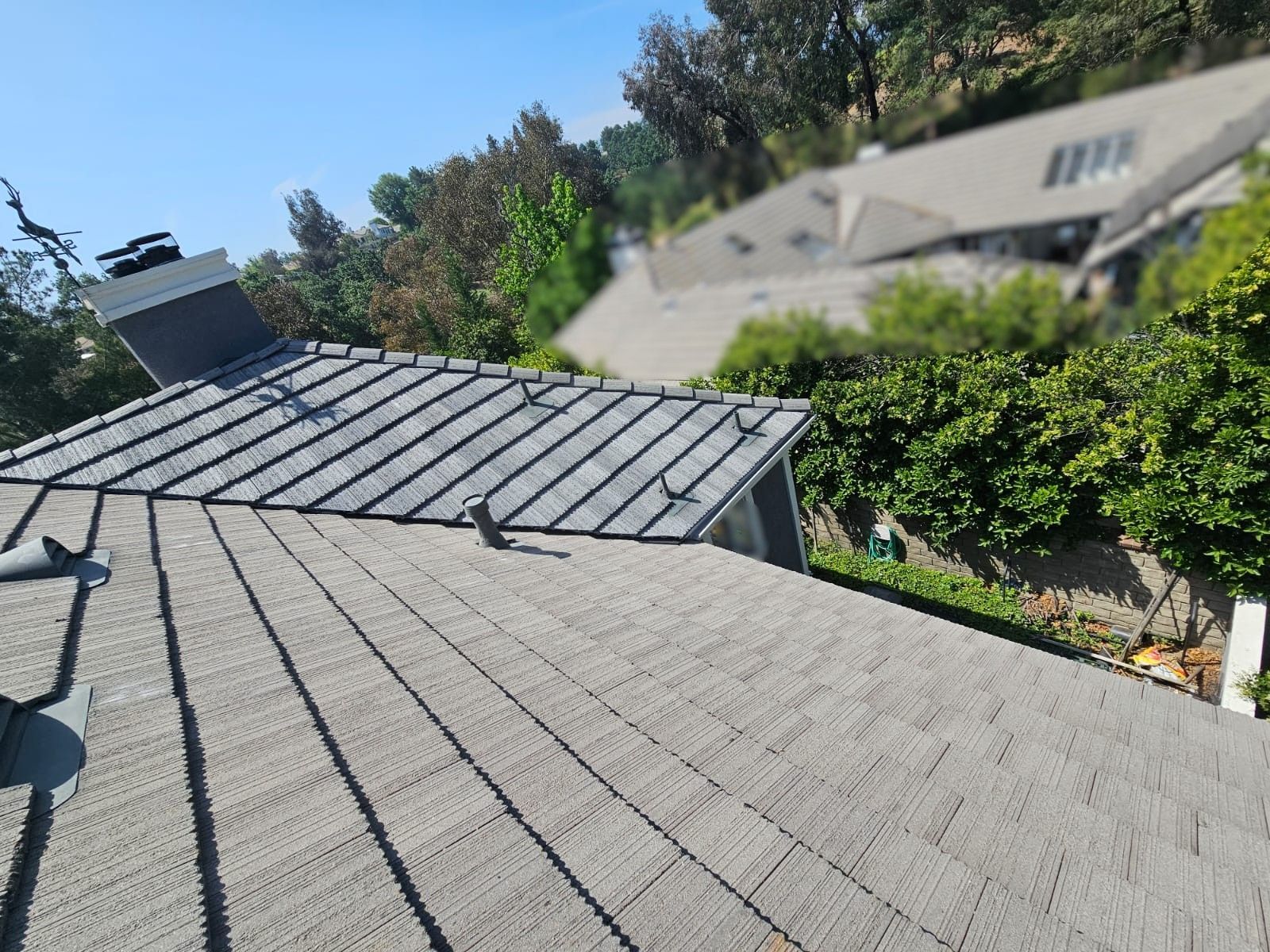 Gray tiled roof of a house with a chimney, trees and a partly visible house in the background on a sunny day.