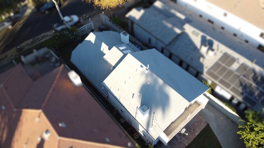 Aerial view of a white-roofed house with a driveway and adjacent buildings, bathed in sunlight.