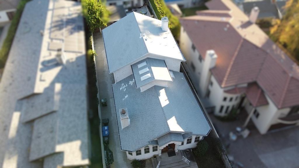 Aerial view of a light gray roofed house, flanked by buildings and trees. Sunny day.
