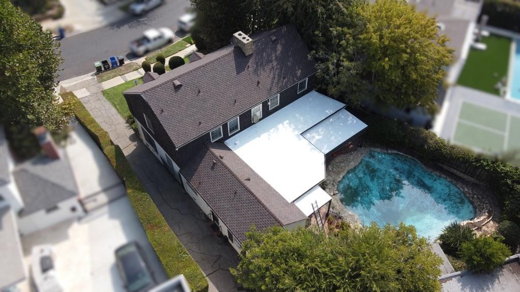 Aerial view of a house with a pool, tennis court, and cars on the street. Dark roof and blue water.