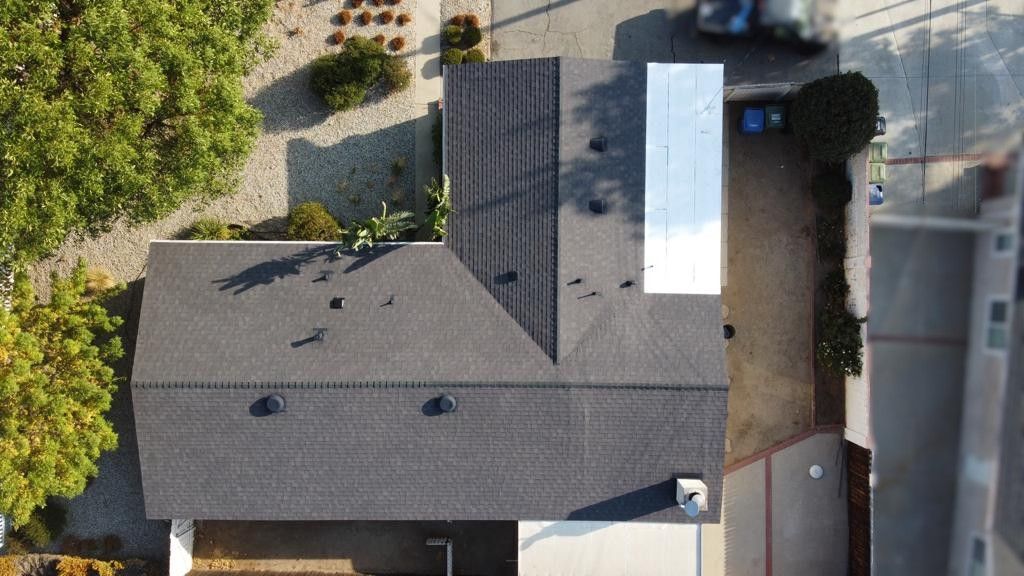 Overhead view of a dark gray shingled house roof with some trees and a car.