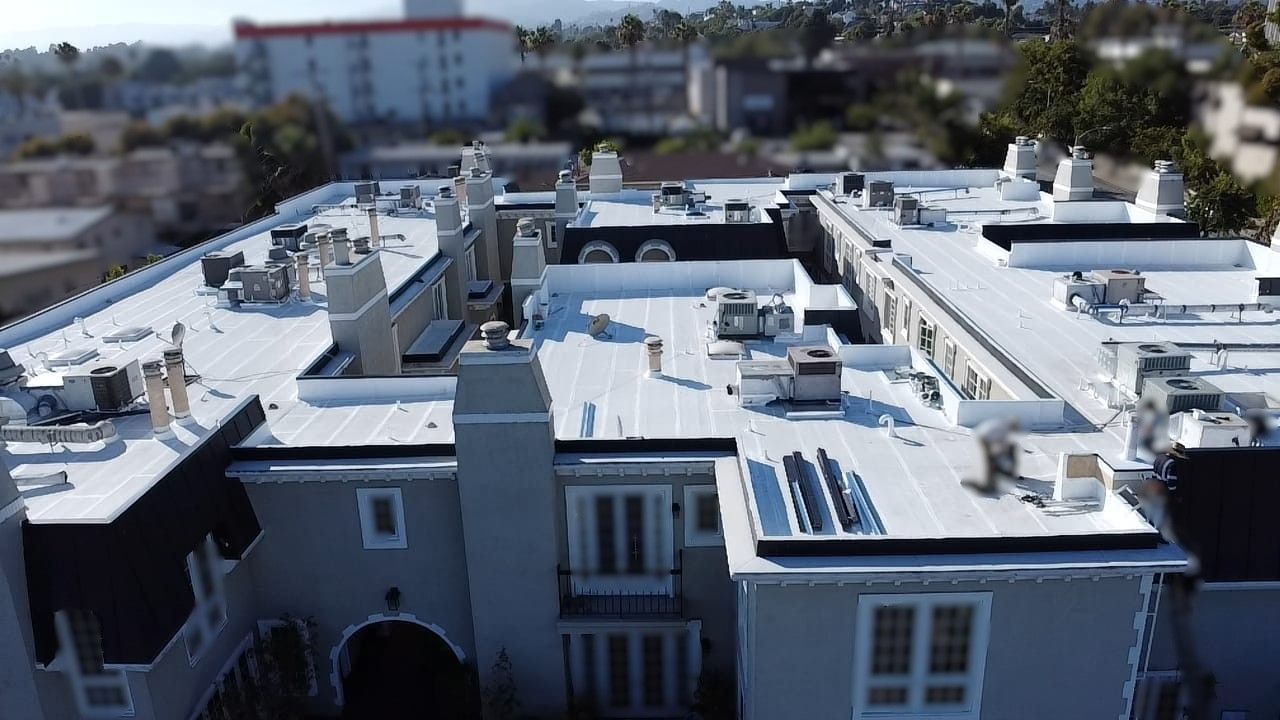 Aerial view of a white-roofed building with chimneys and HVAC units. Other buildings are in the background.
