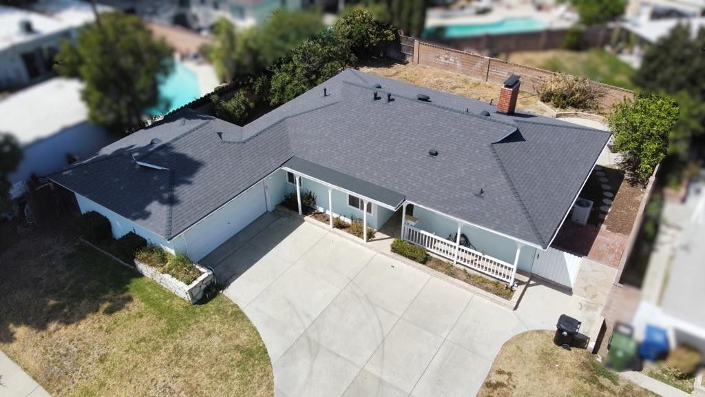 Aerial view of a house with a gray roof, white exterior, driveway, and surrounding green grass and trees.