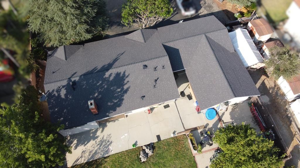 Aerial view of a dark roofed house with a concrete patio, a backyard, and surrounding trees.