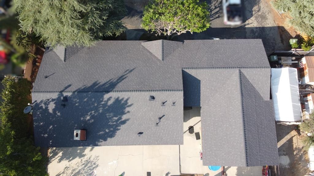 Aerial view of a house with a gray shingle roof, surrounded by trees and a driveway with a white car.