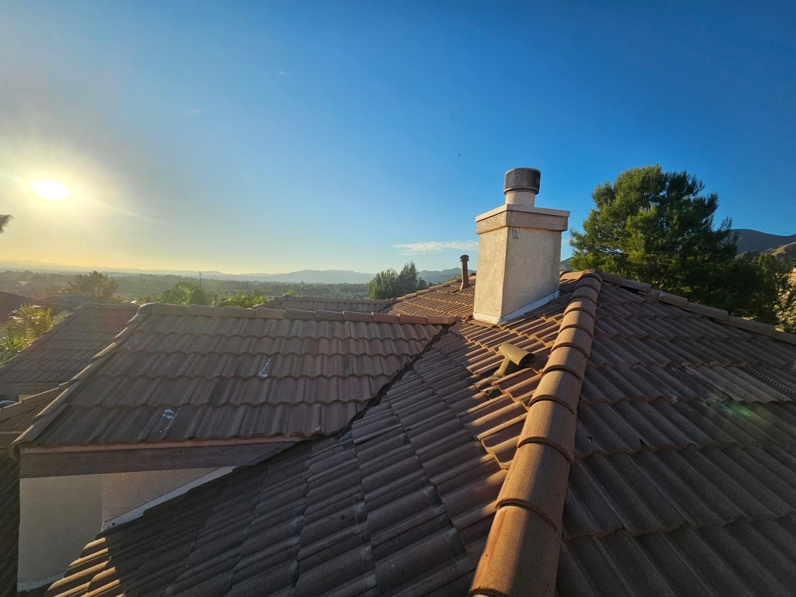 View of rooftops with a chimney against a bright blue sky, sun in the distance.