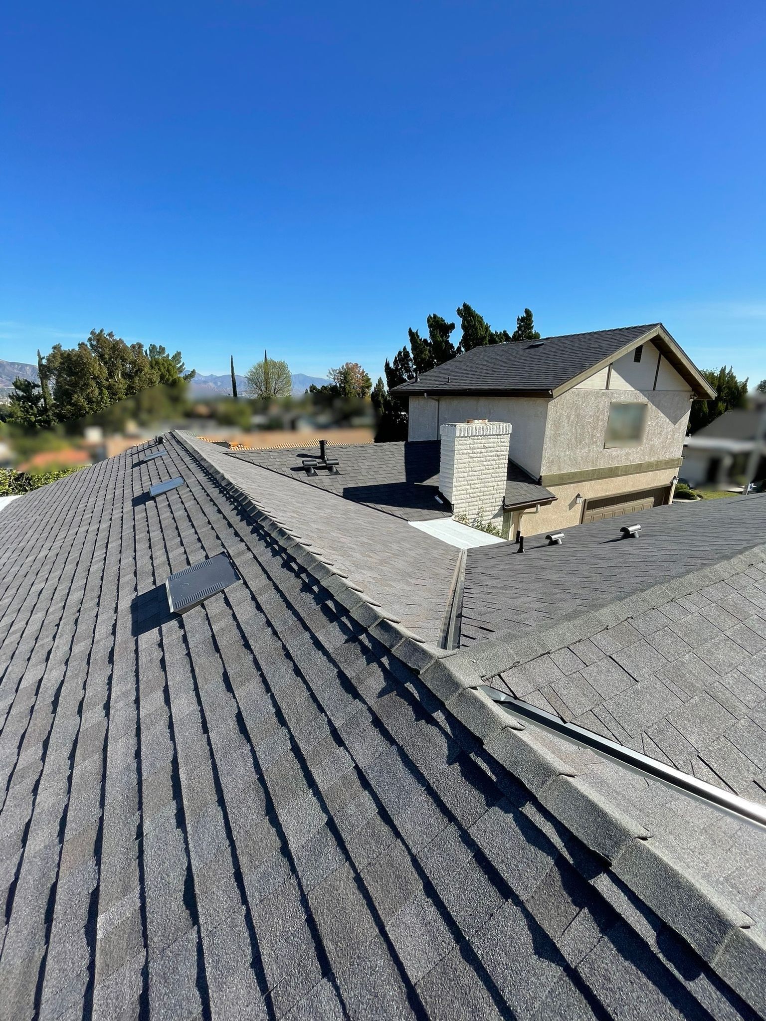 Roof of a house with gray shingles under a clear, blue sky.  A second structure is visible in the distance.