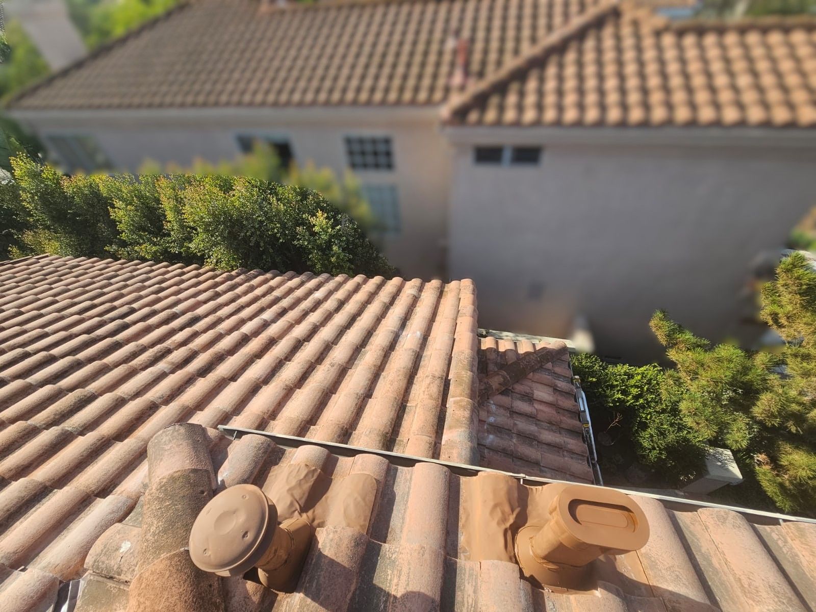 Terracotta tile roof with vents, overlooking a stucco house with green shrubs in the background.
