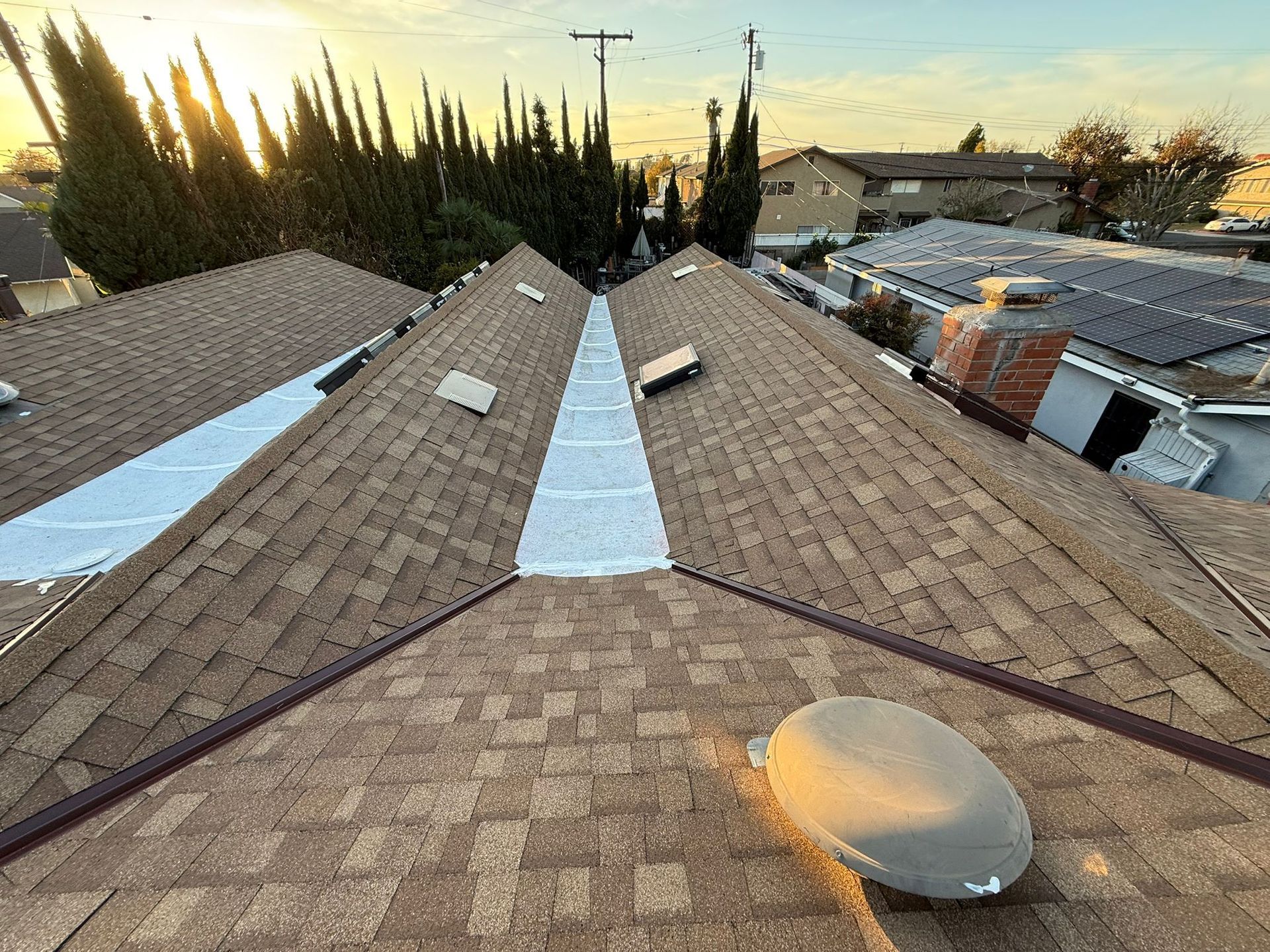 View from rooftop. Brown shingled roof, with white sealant along ridges. Sunset in background.