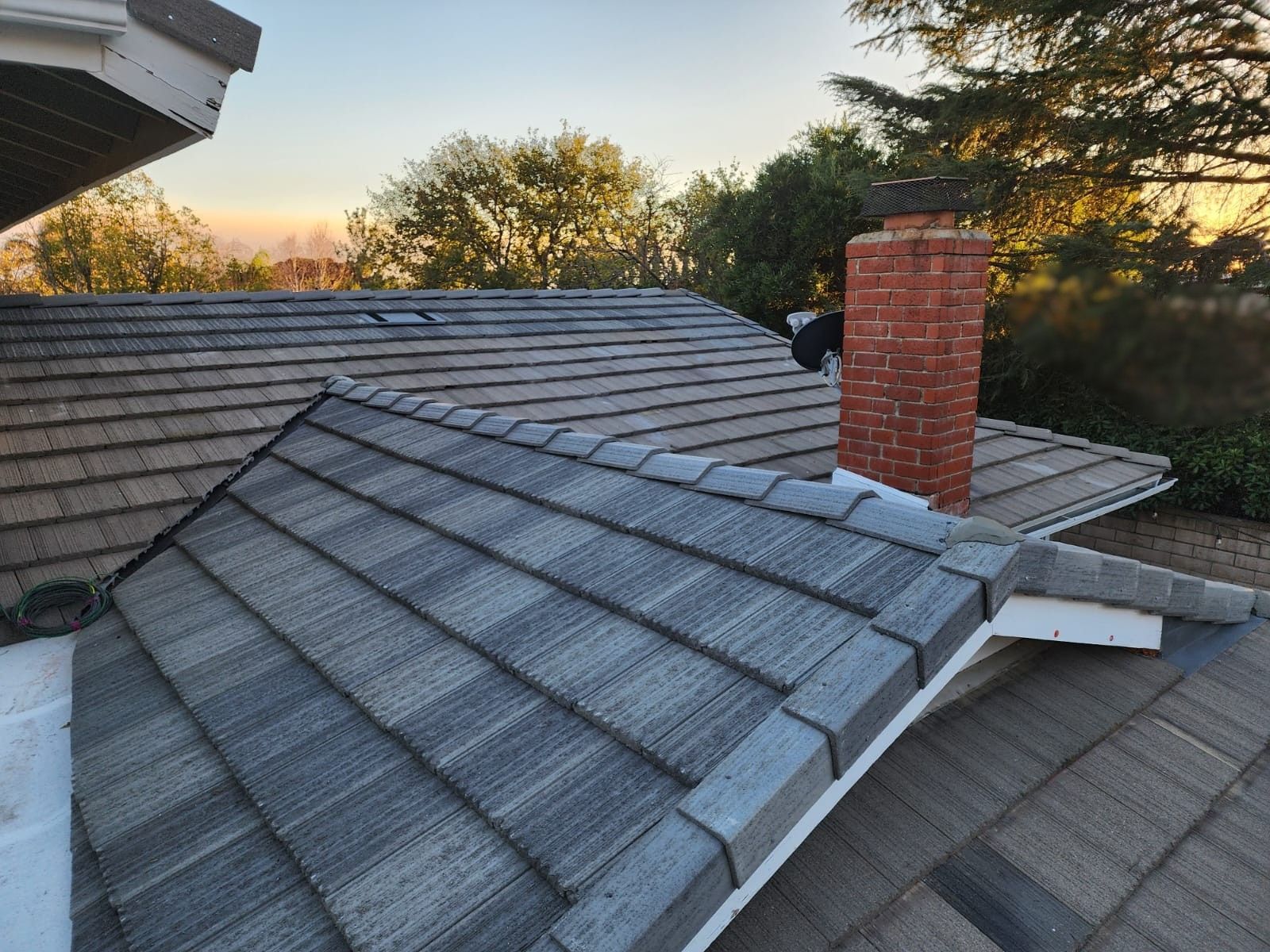 Close-up of house roof with chimney. Gray and brown tiles, brick chimney, tree line backdrop.