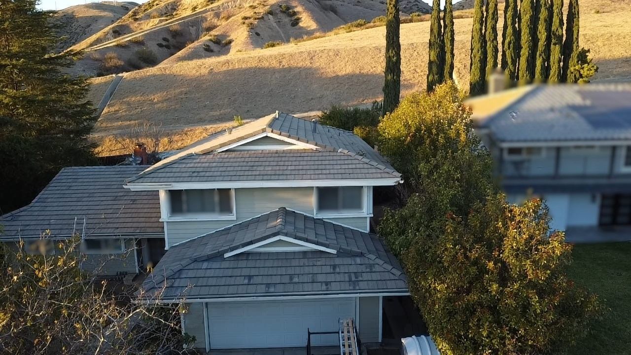 Two-story house with gray roof, green trees, and a hillside in the background.
