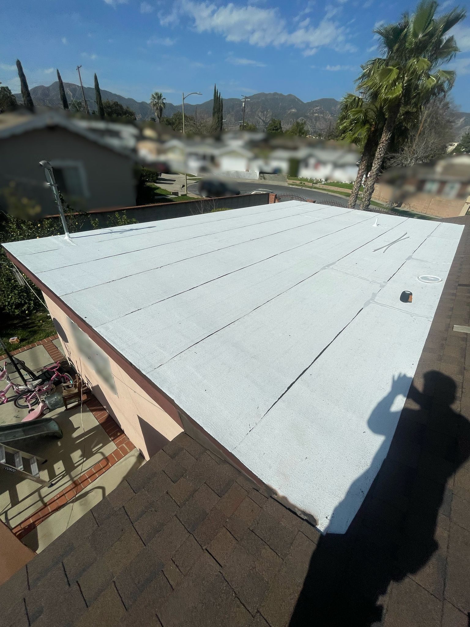 View of a flat white roof being worked on, from an adjacent brown shingled roof. Blue sky, palm tree and mountains in background.