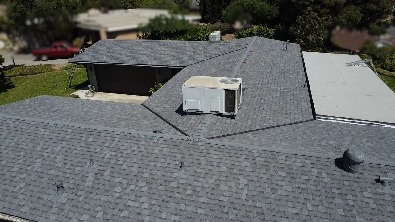 Gray shingled roof with an air conditioning unit, small chimneys, and an out-of-focus background of buildings and trees.