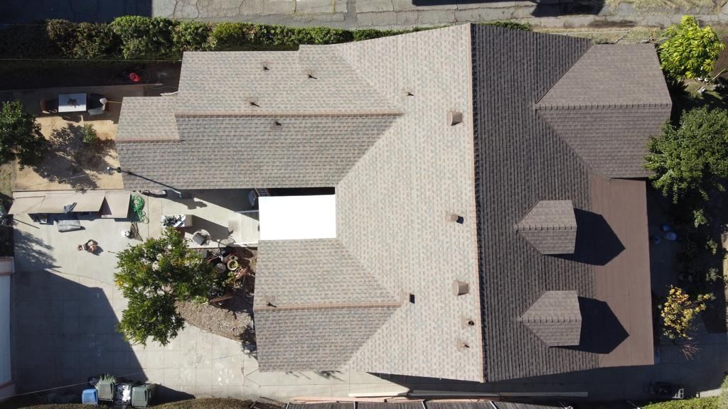 Aerial view of a brown-roofed house with a complex structure, surrounded by trees and parked vehicles.