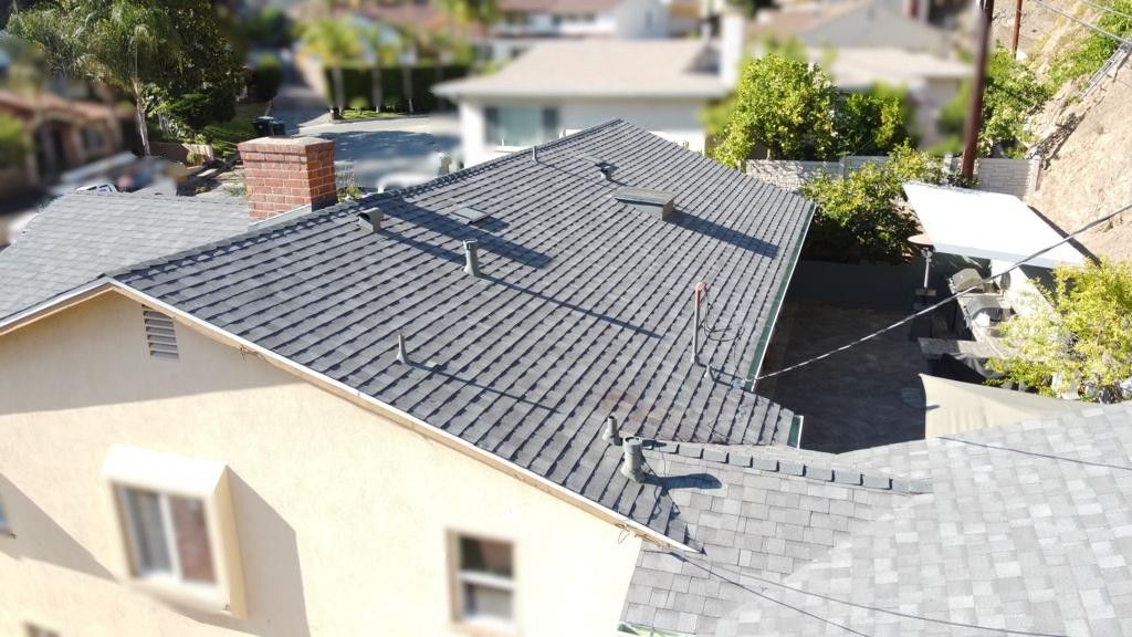 Overhead view of a house with a gray shingled roof, brick chimney, and surrounding homes in a sunny setting.