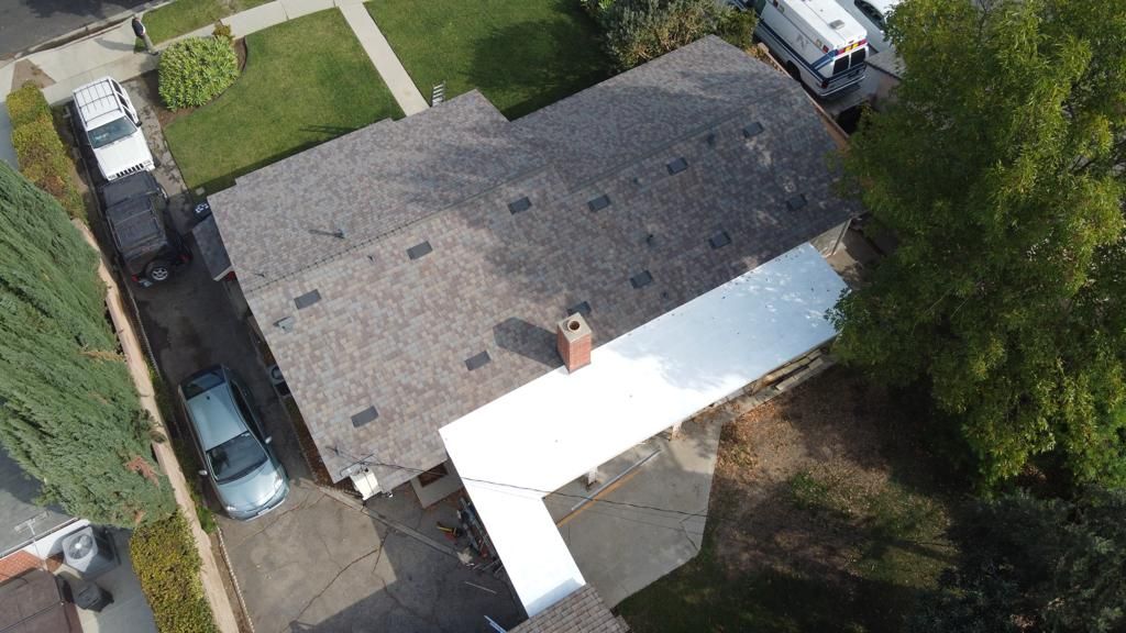 Overhead view of a house with a gray roof and white carport; a van and cars are parked in the driveway.