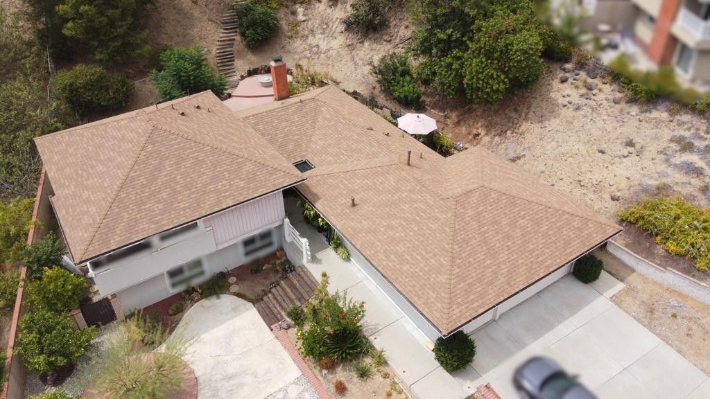 Aerial view of a beige house with brown roof and a driveway, surrounded by trees and shrubs on a hillside.