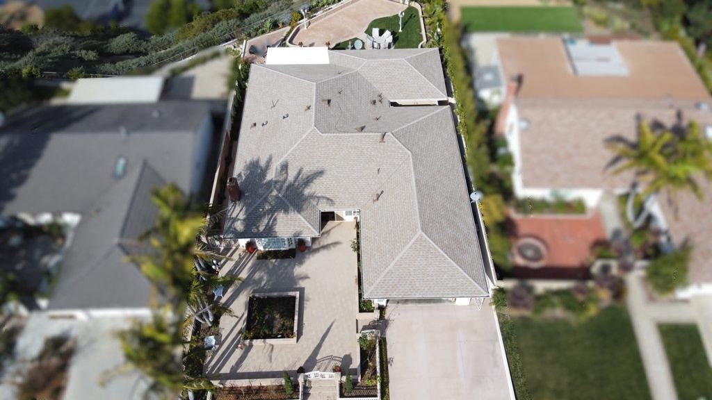 Aerial view of a house with a gray roof and a long driveway, surrounded by greenery and other houses.