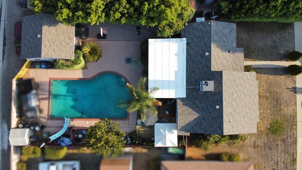 Aerial view of a house with a pool, patio, and solar panels, surrounded by trees and other houses.