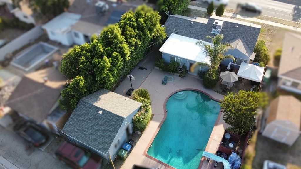Aerial view of a house with a pool, surrounded by trees and other houses on a sunny day.