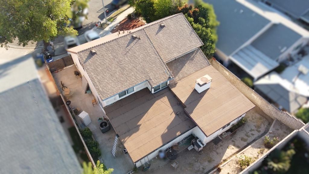 Aerial view of a house with brown roof and deck, white walls, surrounded by a fence and some trees.