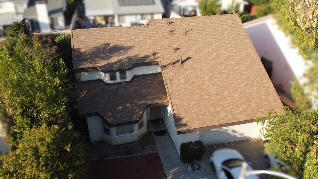 Aerial view of a house with a brown roof, white walls, and a car parked in the driveway, surrounded by trees.