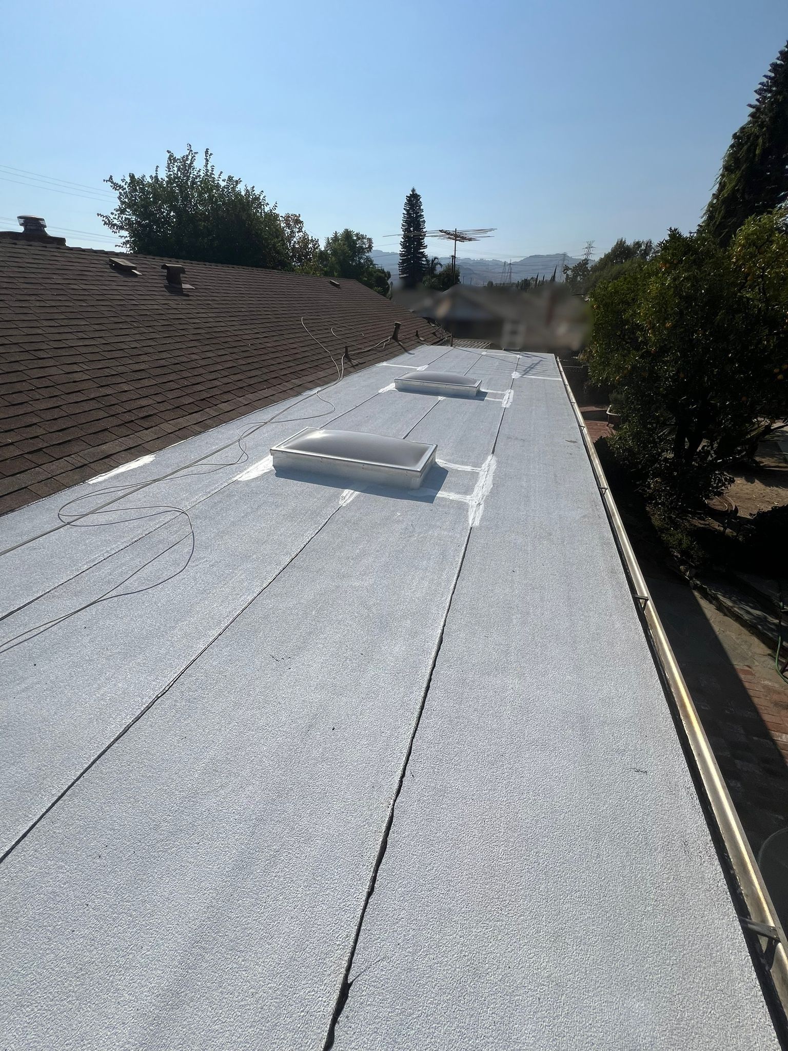 A newly-installed flat roof with skylights on a sunny day. Adjacent is a brown shingled roof.