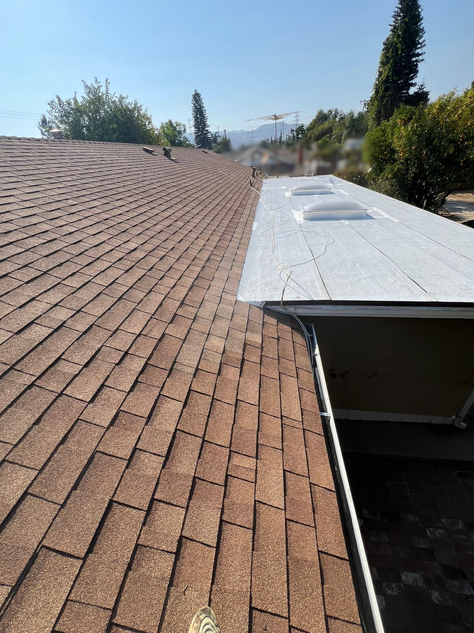 A brown shingle roof meets a white flat roof with skylights, under a clear blue sky.