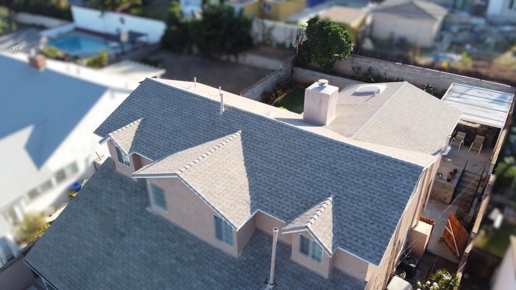 Aerial view of a house with a brown roof and beige exterior. Other houses and a pool are visible in the background.