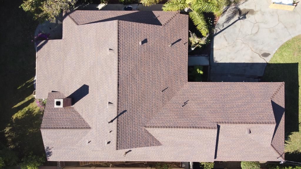 Aerial view of a brown-roofed house with a chimney, driveway, and surrounding green lawn.