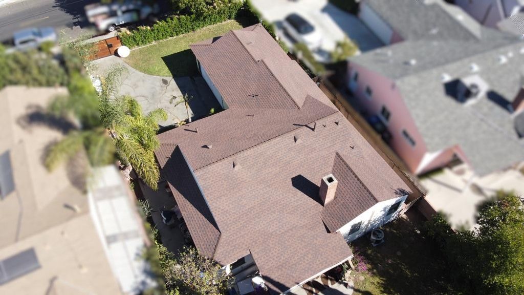 Overhead view of a brown-roofed house with a chimney, surrounded by green trees and other houses.