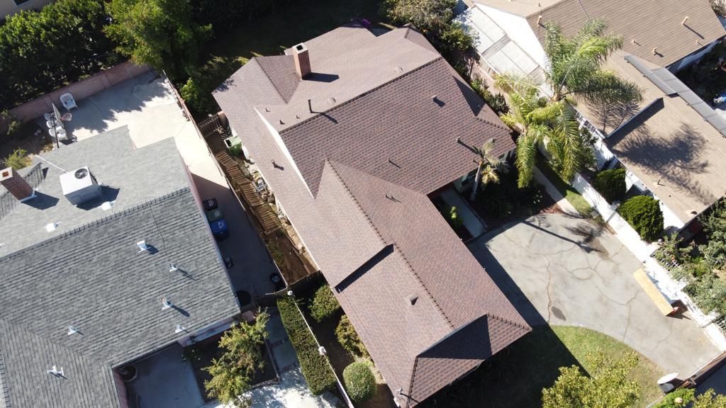 Overhead view of a brown-roofed house, with a chimney, and a driveway; located between other houses with trees.