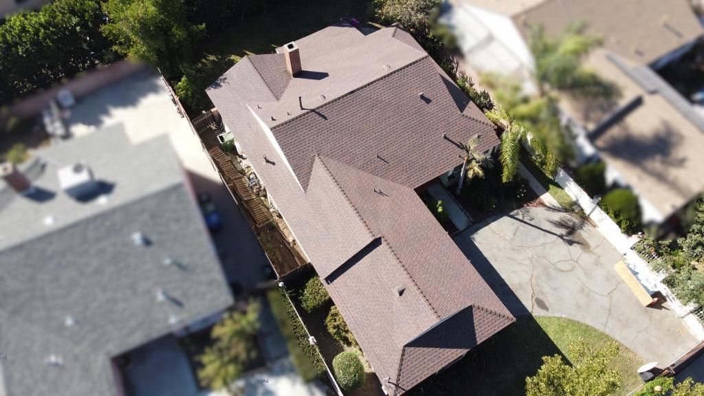 Overhead view of a two-story house with a brown roof and driveway, surrounded by trees and other houses.