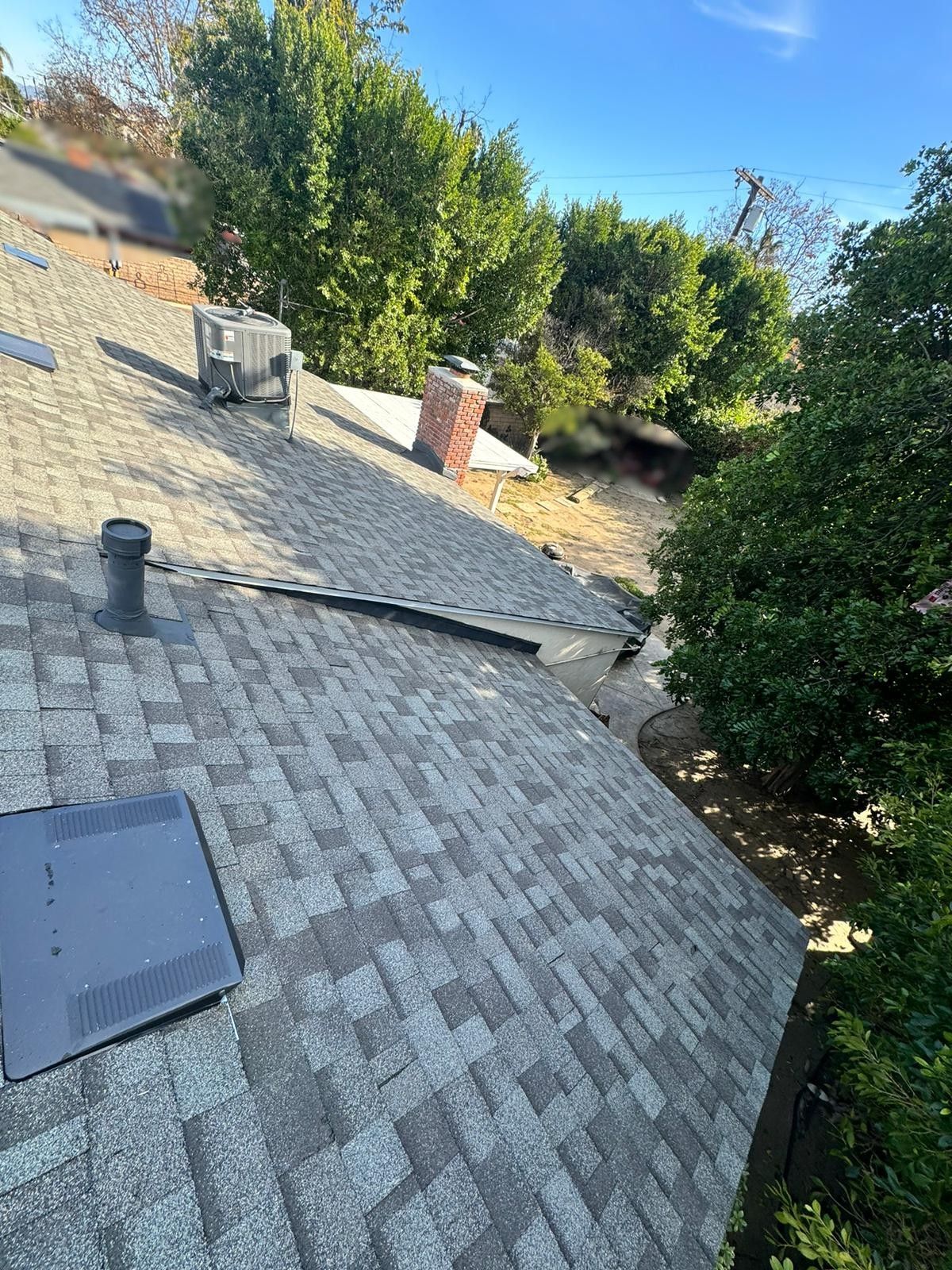 Roof with shingles, vent pipes, and a brick chimney, surrounded by trees and a blue sky.