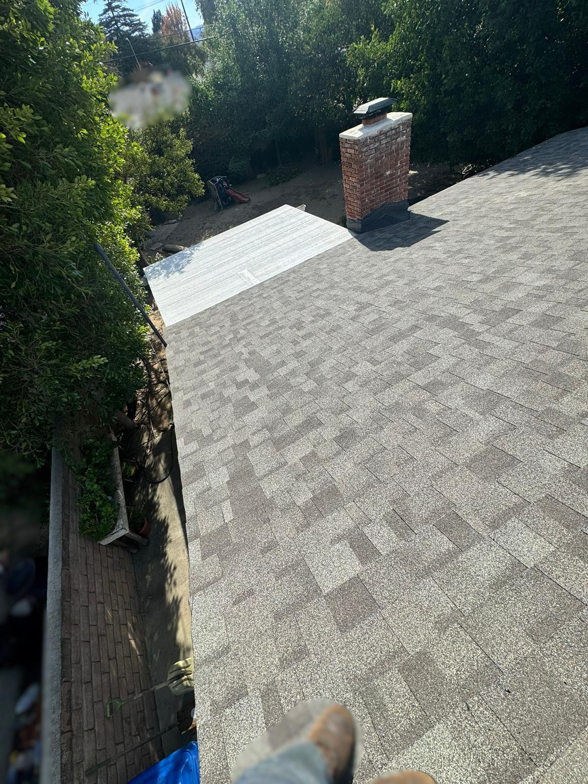 A roof with gray shingles, a chimney, and a metal sheet area. Trees and greenery are in the background.