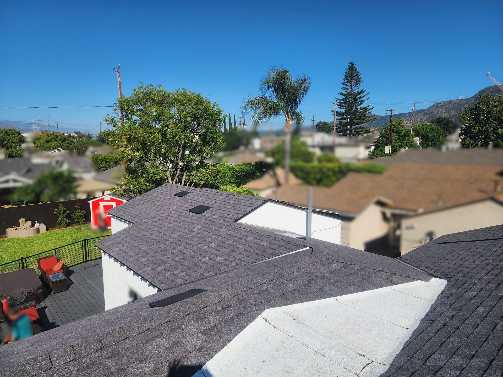 Rooftop view of neighborhood homes with gray shingles, green trees, and a bright blue sky.