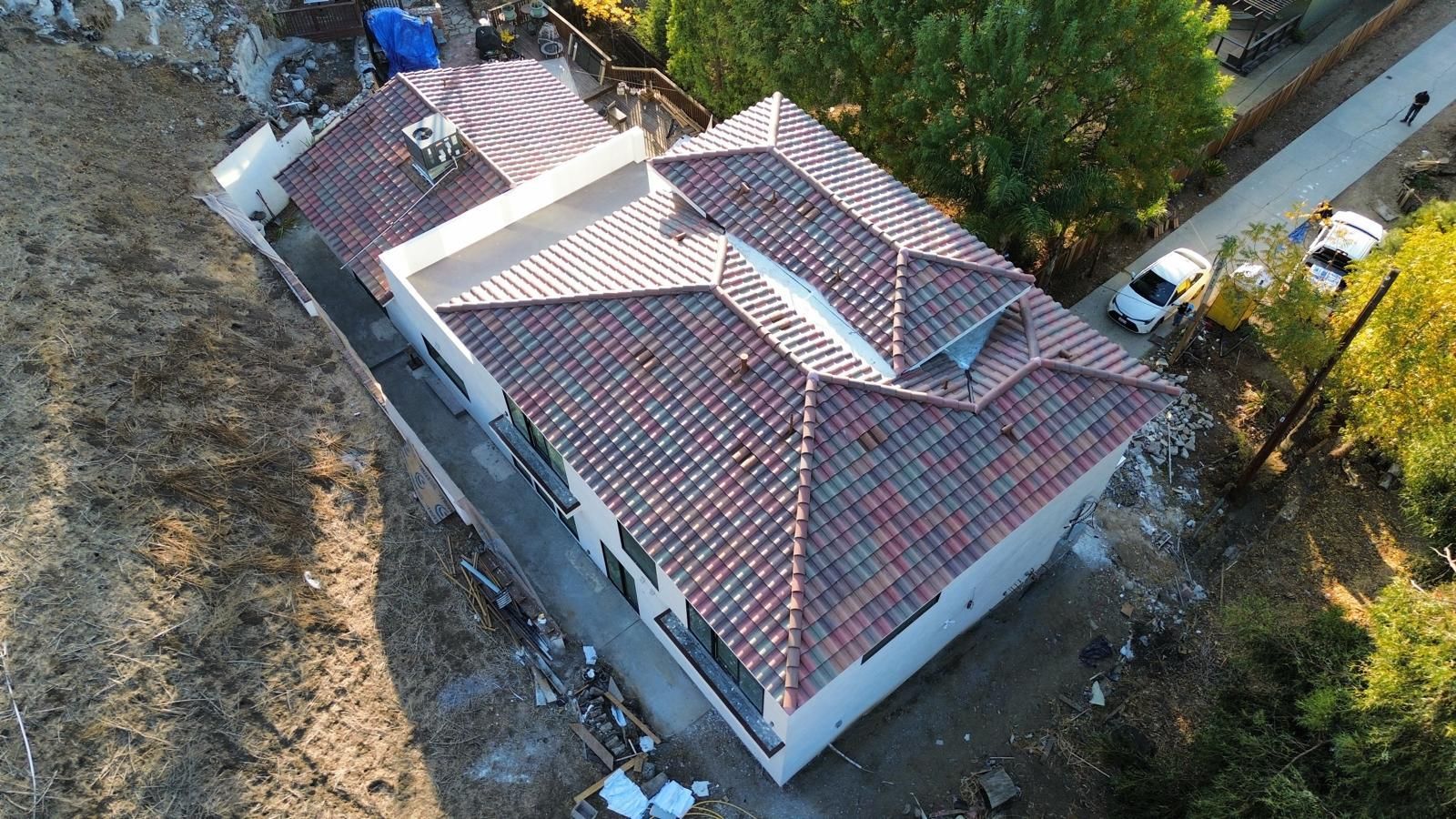 Aerial view of a house with a red tile roof, surrounded by dry land and trees; cars parked nearby.